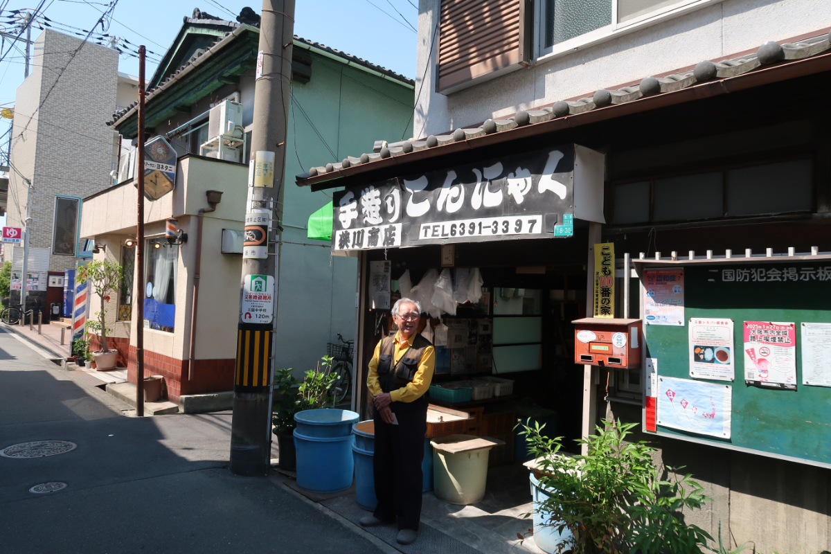 大正13年築(102年前)の狭川商店は入母屋作りの銅板張りです。「手作りこんにゃく」の黒い看板も狭川さんの手作りです。お隣の床屋も狭川商店と同じ作りで狭川さんの祖父の代から続いています。昔はこの通りも賑やかな商店街だったそうです。戦中には近くの「日本アルミ」工場を狙い爆撃があったそうですが、爆弾は北に逸れて狭川商店の一角は焼け残ったそうです