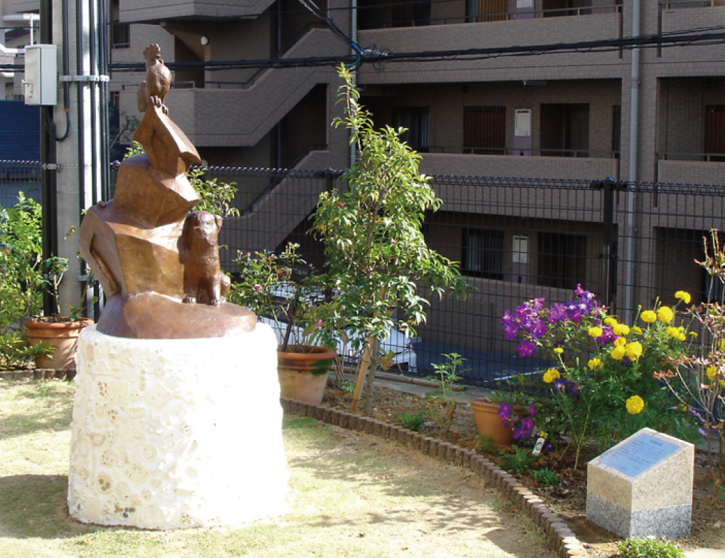 《ひだまりが丘-春》 ブロンズ 吹田市　特別養護老人ホーム　ちくりんの里 – 犬と猫と鶏、バラバラな３匹が家族になる。台座部分に埋め込まれた陶板レリーフは、ホームの入居者や関係者と共に制作した。「作家がものをつくっておしまいでは面白くないと思って。とにかく全員参加！　と言ったらみんな一丸となって盛り上がってくれて、楽しい仕事でしたね」